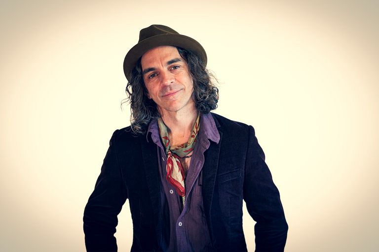 Australian blues musician Nathan Cavaleri smiling in a fedora, dark jacket, purple shirt, and patterned neck scarf against a plain backdrop.